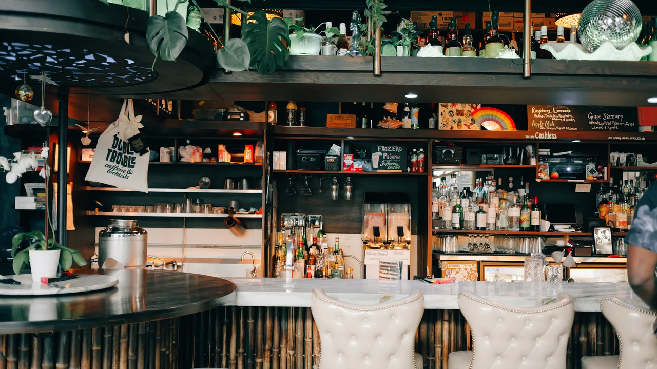 An eclectic bar interior is filled with bottles, objects, and worn surfaces.